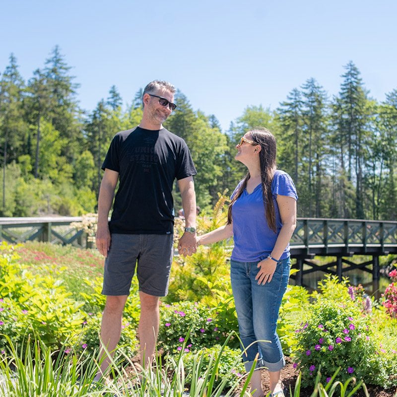 Couple standing in a garden in summer clothes and looking at each other with pine trees behind them in the distance.