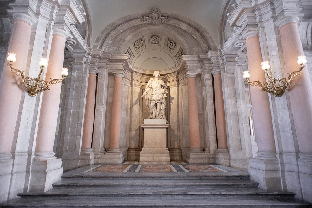 Sculpture of Charles III in Spain at the entrance of the Royal Palace of Madrid in front of a marble wall, surrounded by pink columns and gold sconces.