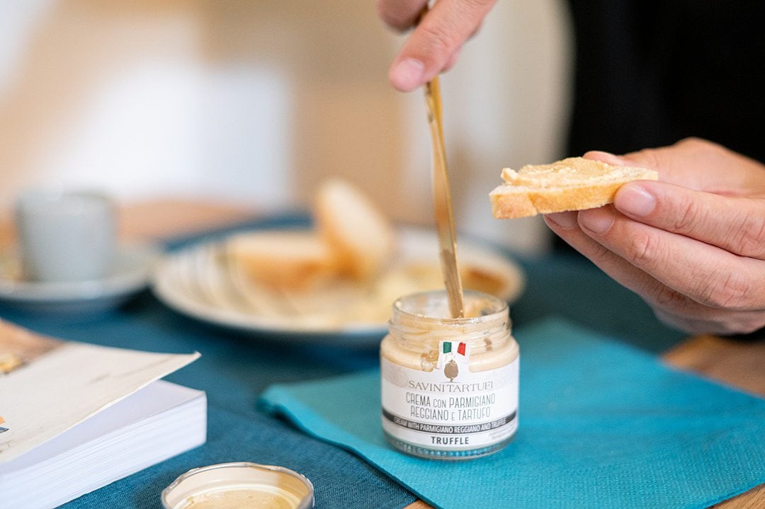 Knife dipping into a jar of truffle cheese spread on a blue napkin, next to a hand holding a slice of bread.