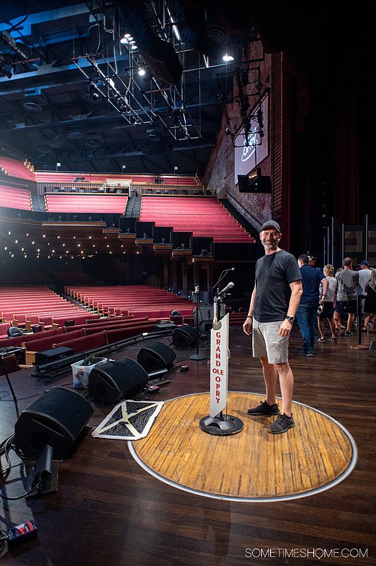 Man standing on stage, on the famous wood floor circle at the Grand Ole Opry with a microphone stand in front of him.
