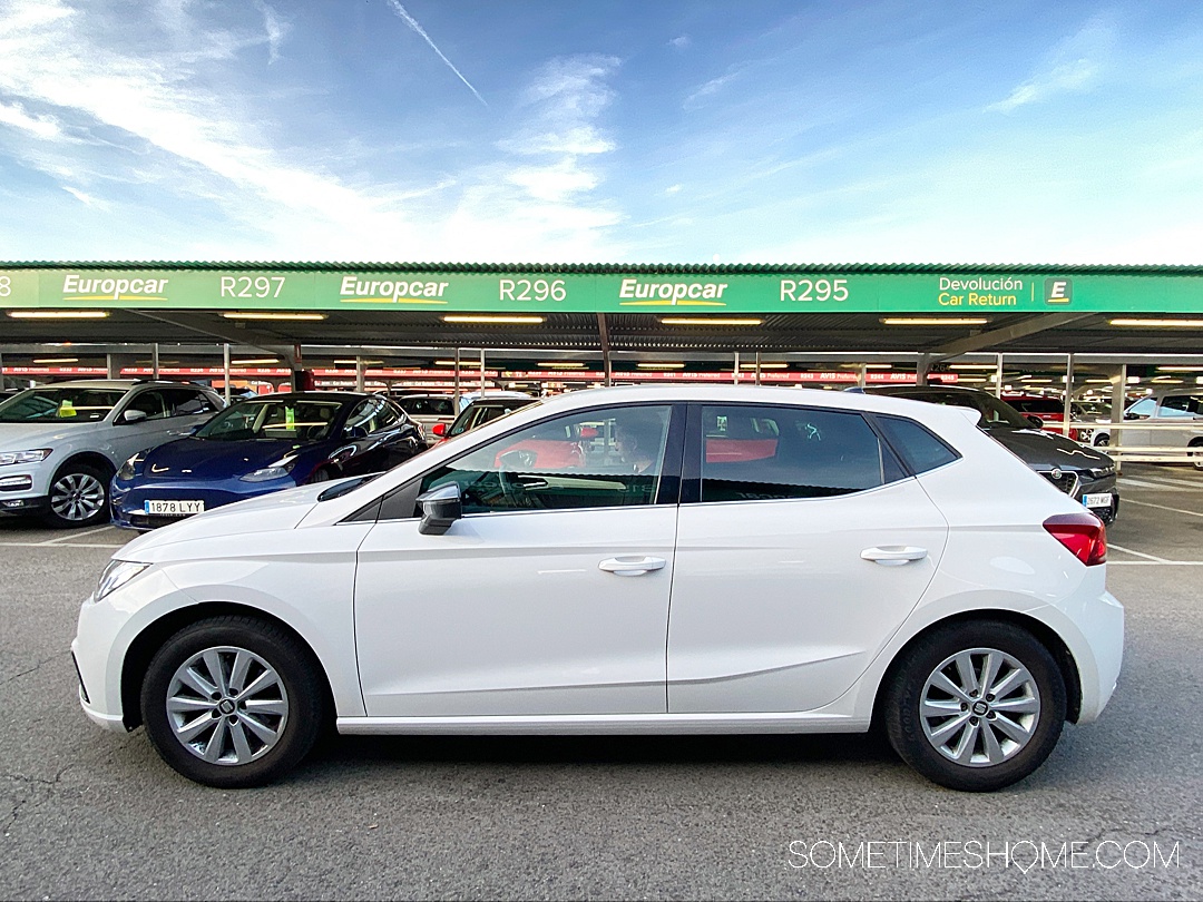 White car profile in a parking lot with a green sign overhead renting a car in Madrid.