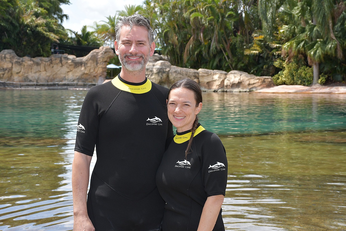 Man and woman in short sleeve wetsuits with a lagoon behind them at Discovery Cove theme park in Orlando, FL.