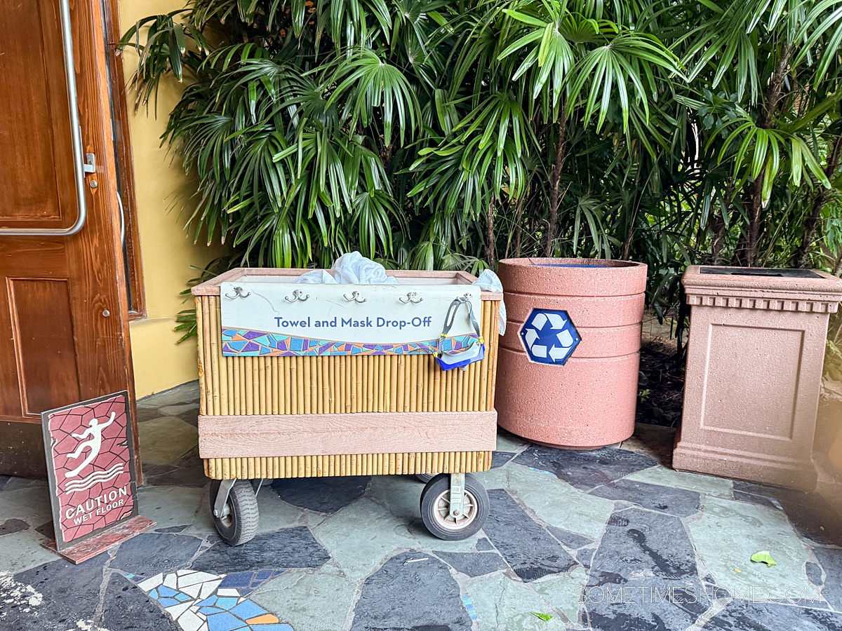 Bin to dispose of towels and snorkel masks next to a recycling bin, in front of tropical leaves.
