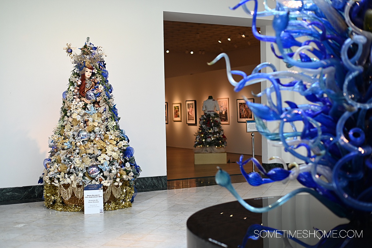 Glass Chihuly sculpture in the foreground on the right with a Christmas Tree on the left in the distance decorated in gold and blue.
