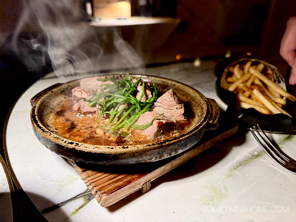 Sizzling meat on a hot dish with seaweed green on top and a plate of French fries next to it. 