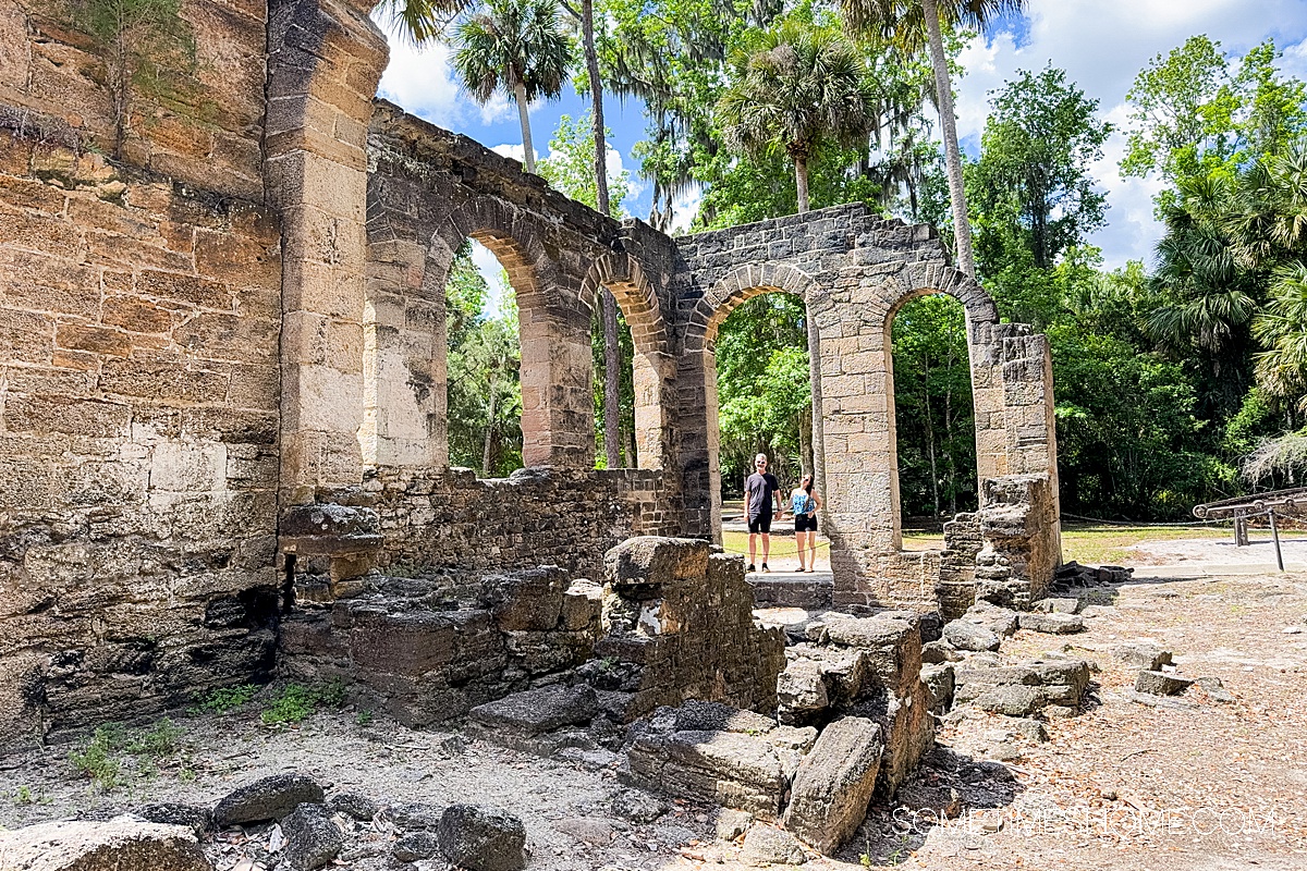 Sugar mill ruins and palm trees surrounding it with a couple seen through an archway in the distance.