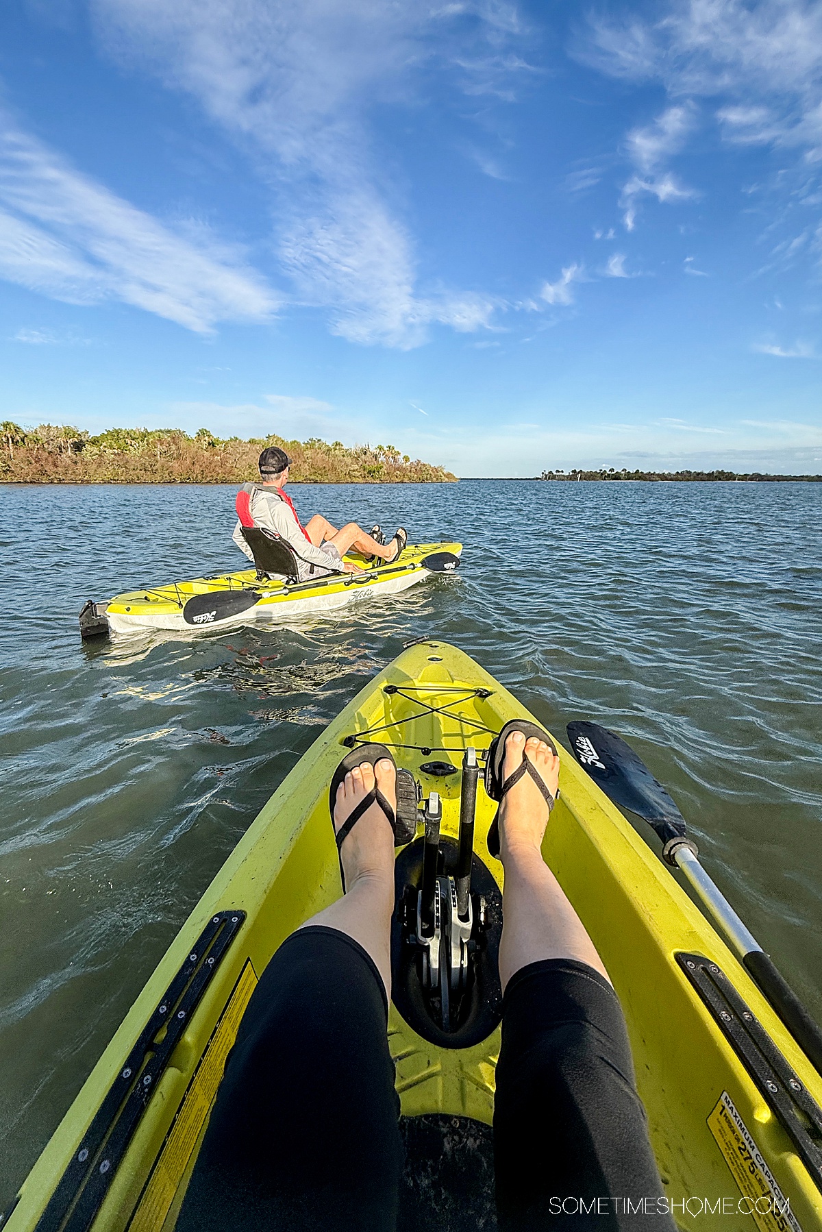 The front of a kayak with feet pedaling and a man ahead in his kayak on the water.